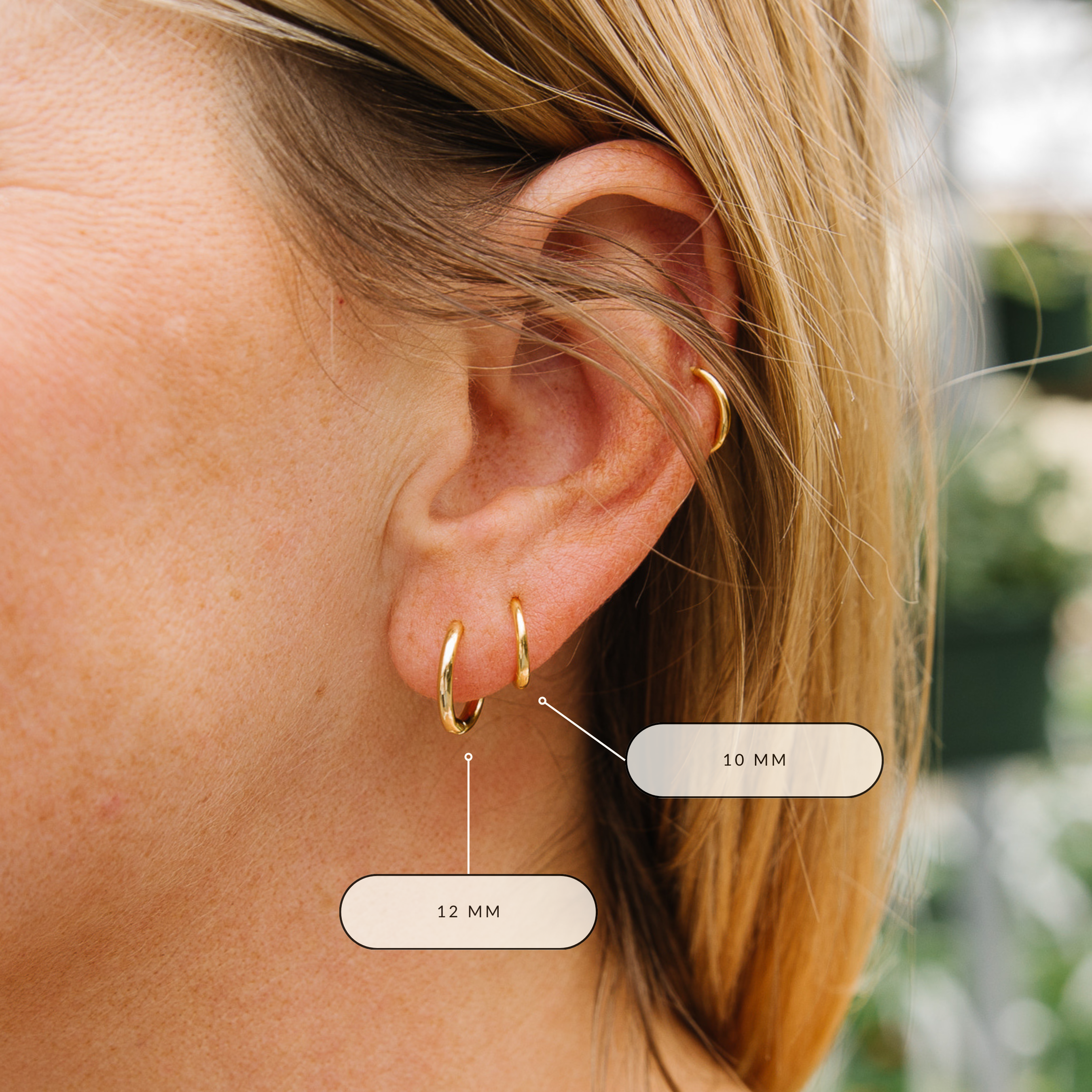 Close-up of a person wearing gold hoop earrings with size labels. The 12 mm gold hoop is in first piercing. 10 mm gold hoop is in second piercing.