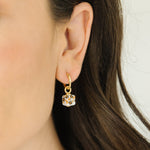 A close-up of a model's ear wearing a delicate gold huggie hoop earring with a small dangling box charm with a black and orange floral design.