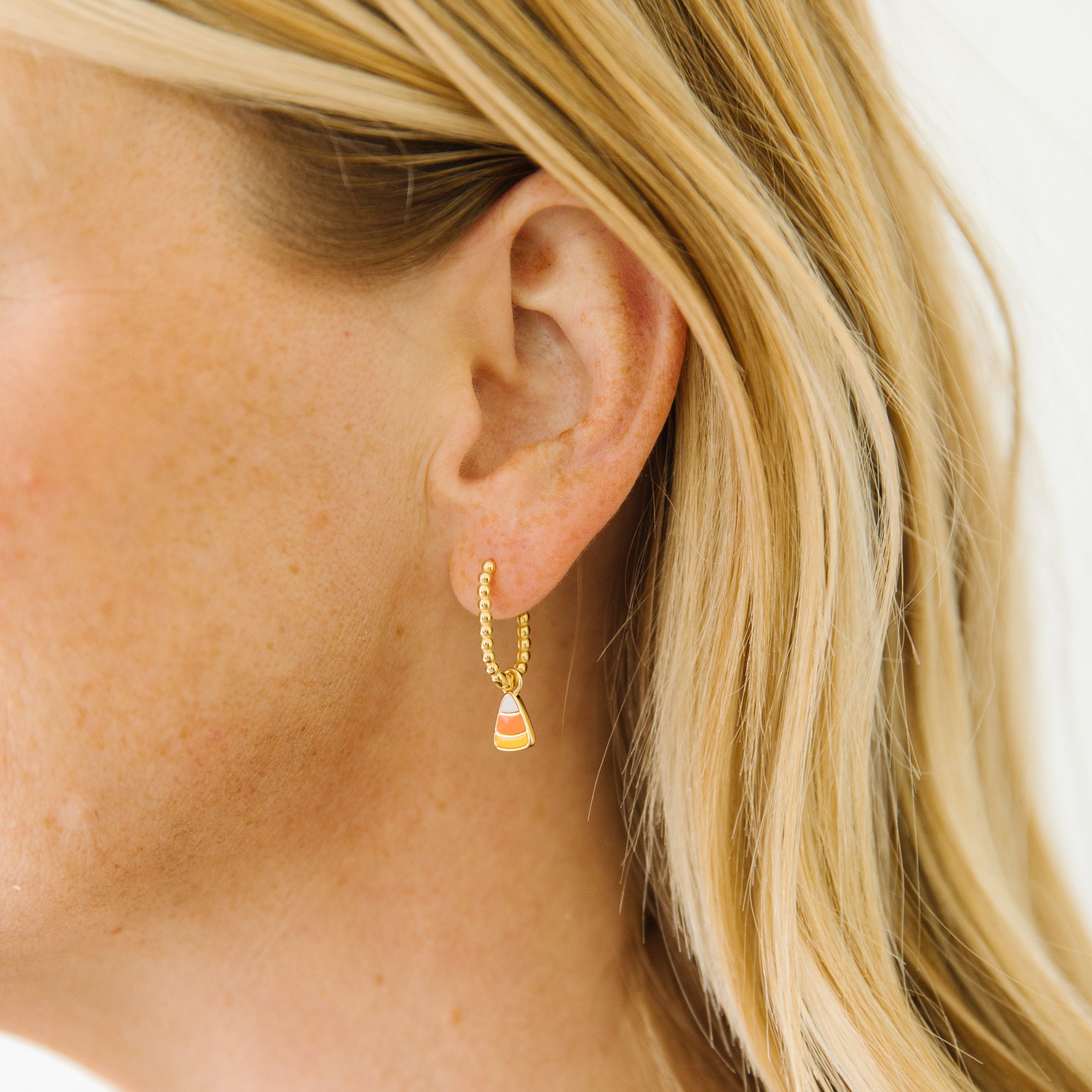 A close-up side profile of a woman with blonde hair wearing a delicate gold huggie hoop earring with a small dangling candy corn charm.
