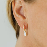 A close-up of a model's ear wearing a delicate gold huggie hoop earring with a small ghost white stone dangle.