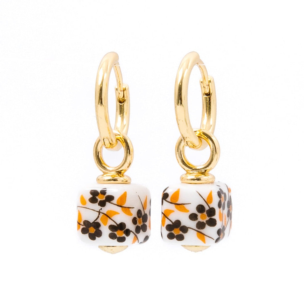 A pair of gold hoop earrings with dangling square charms featuring a white background and a black and orange floral pattern, set against a plain white background.