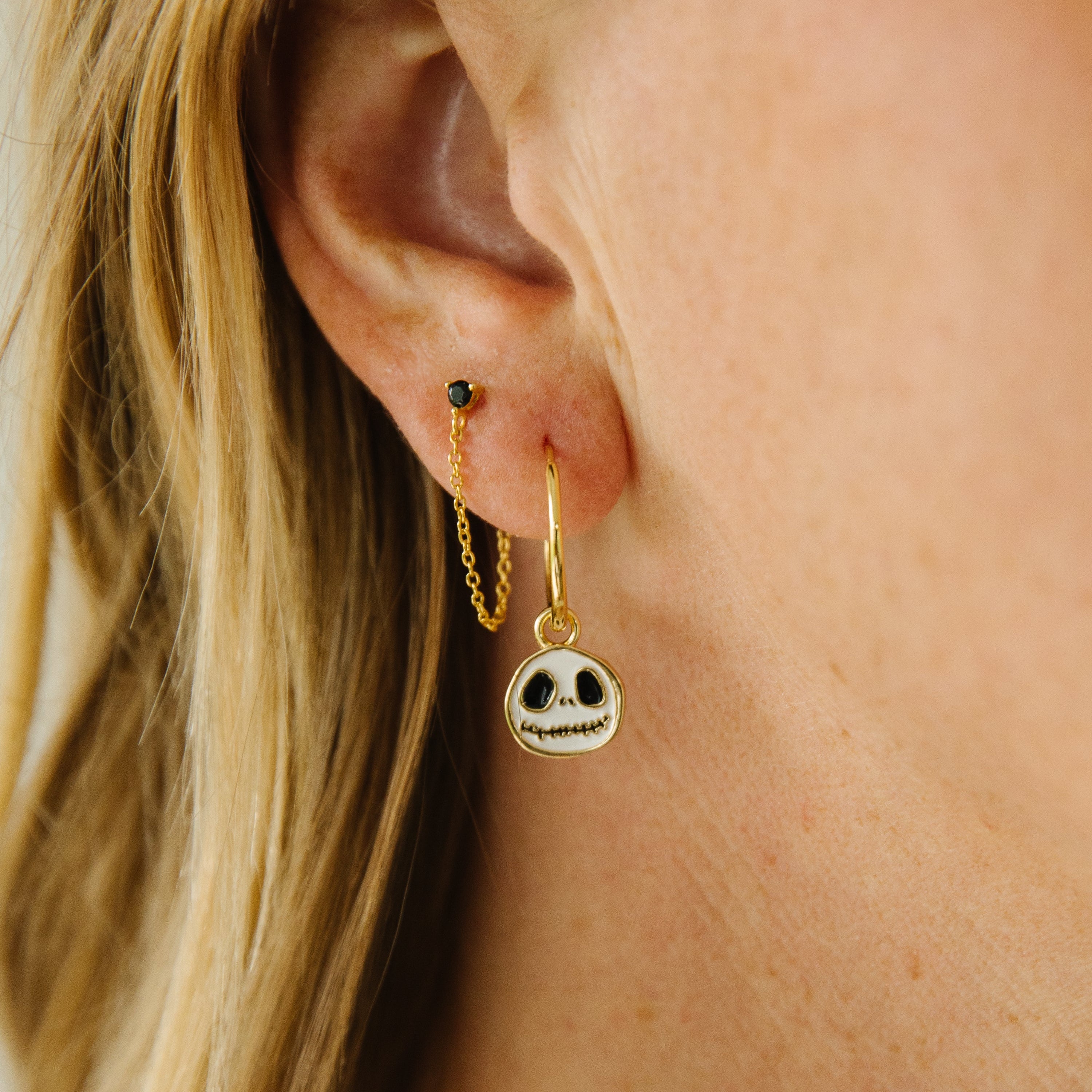 Close-up of a person's ear wearing a gold dangle earring with a small skull charm, alongside a black stud and a thin gold chain.
