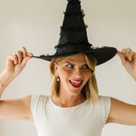 A woman wearing a tall black witch hat and a white sleeveless top, holding the brim of the hat with both hands against a plain light background wearing Jack Skellington charms hanging from gold hoops.