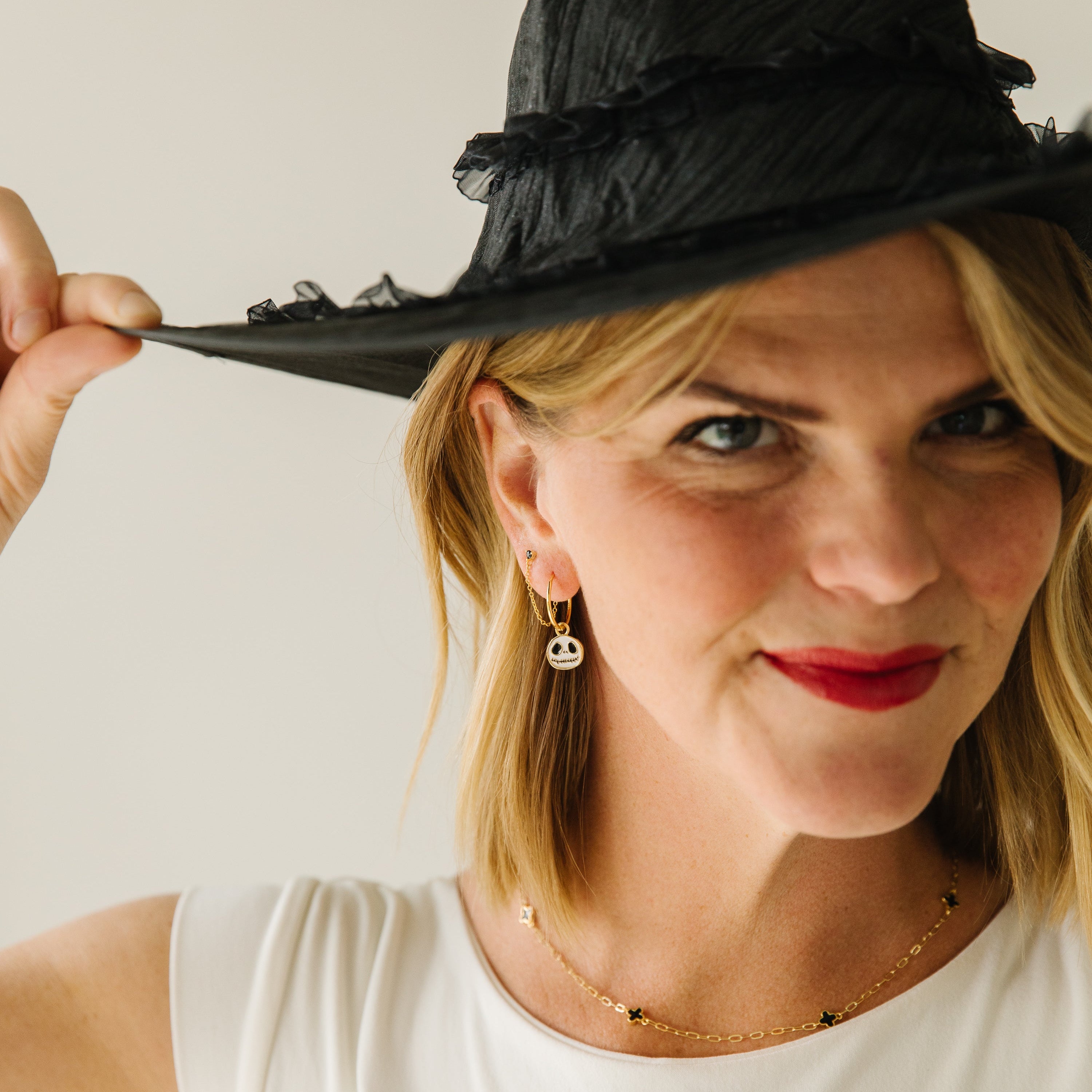 A woman wearing a black ruffled witch hat and a white top, holding the brim of the hat with one hand against a plain light background wearing hoops with  Jack Skellington charms dangling.