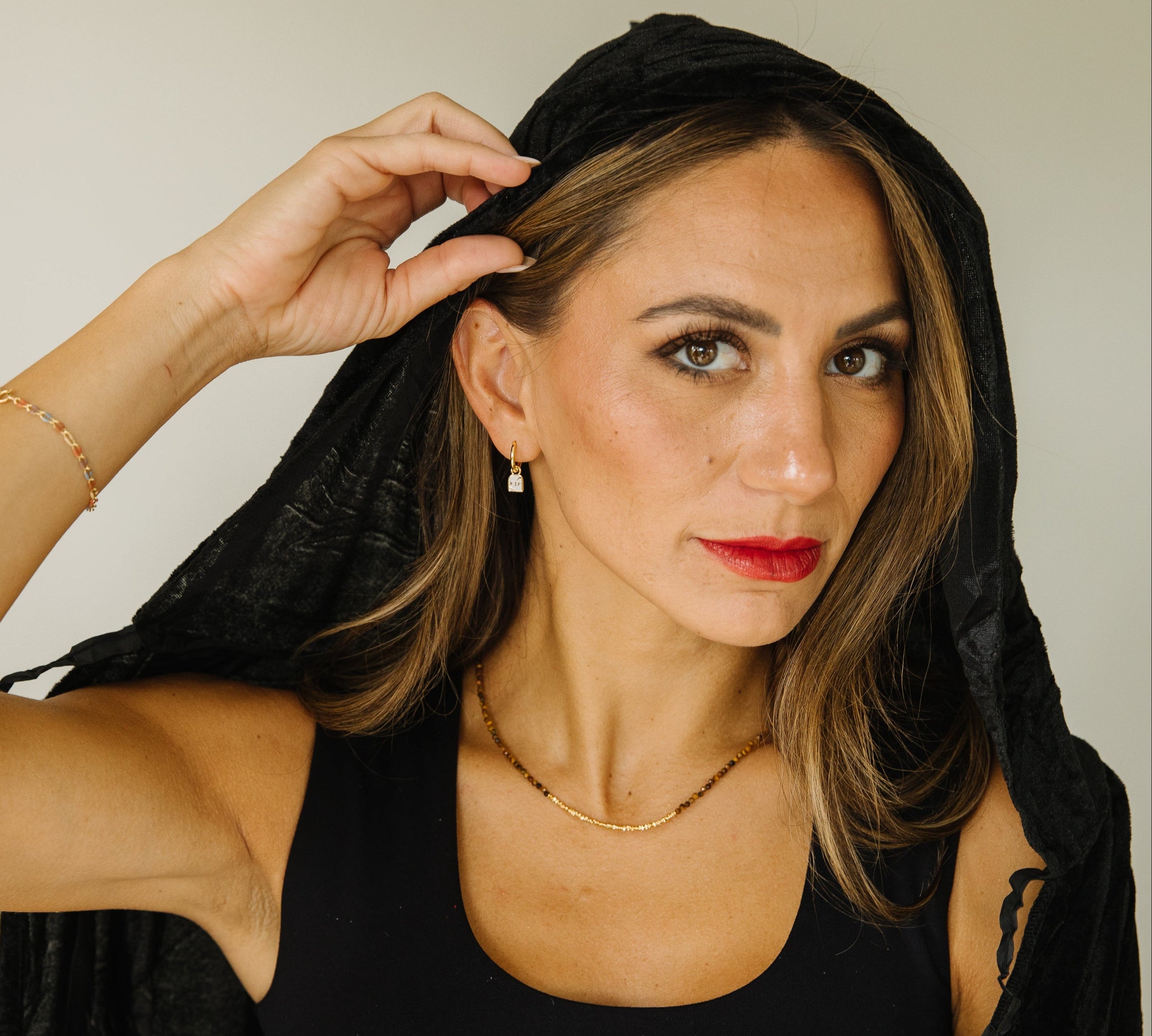 A woman with light brown hair wearing a black sleeveless top and a black hood, posing against a neutral light gray background. She is wearing tombstone earrings.