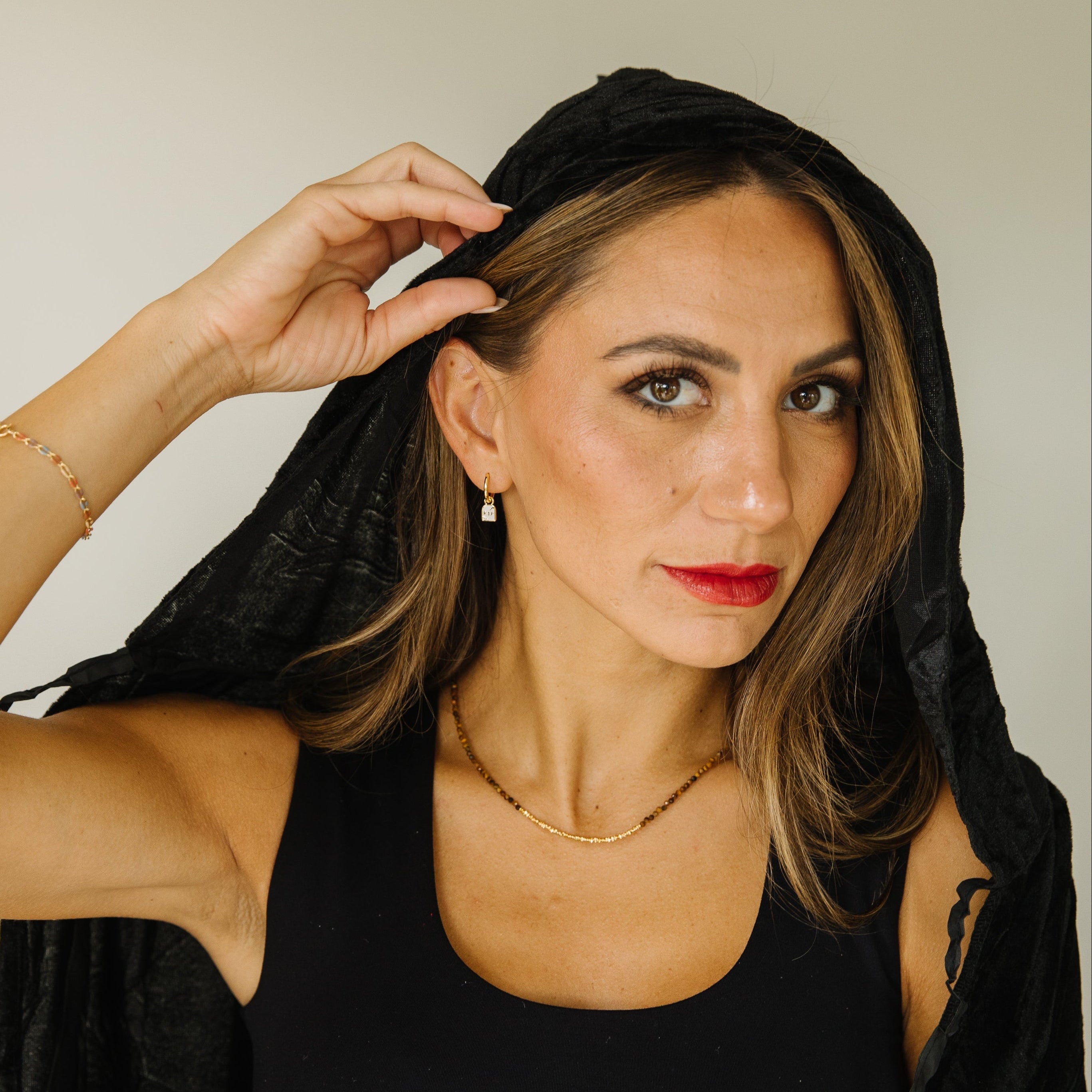 A woman with light brown hair wearing a black sleeveless top and a black hood, posing against a neutral light gray background. She is wearing tombstone earrings.