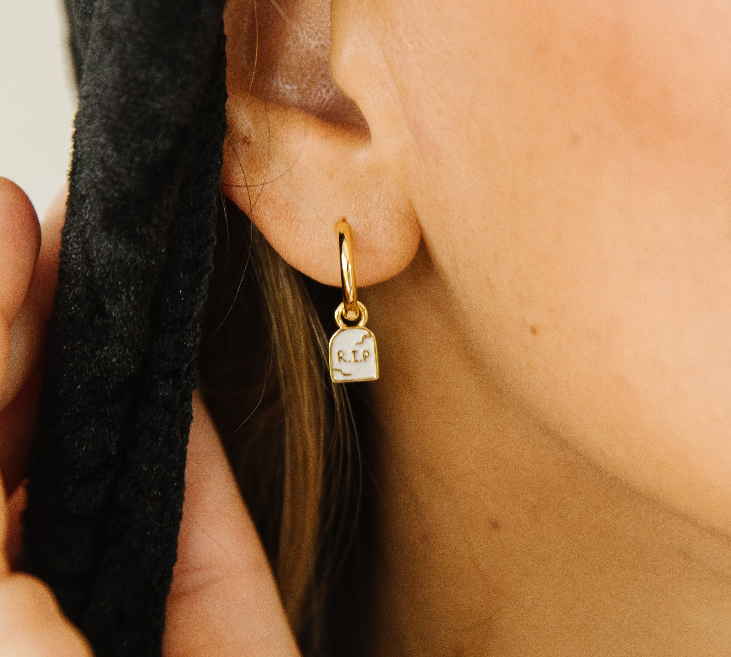 A close-up of a person's ear wearing a small gold hoop earring with a tiny tombstone pendant engraved with RIP.