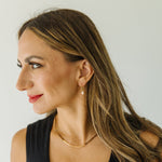 A woman with long brown hair is shown in profile wearing a delicate gold chain necklace and a small gold hoop earring  with light pink ghost charm against a plain light background.