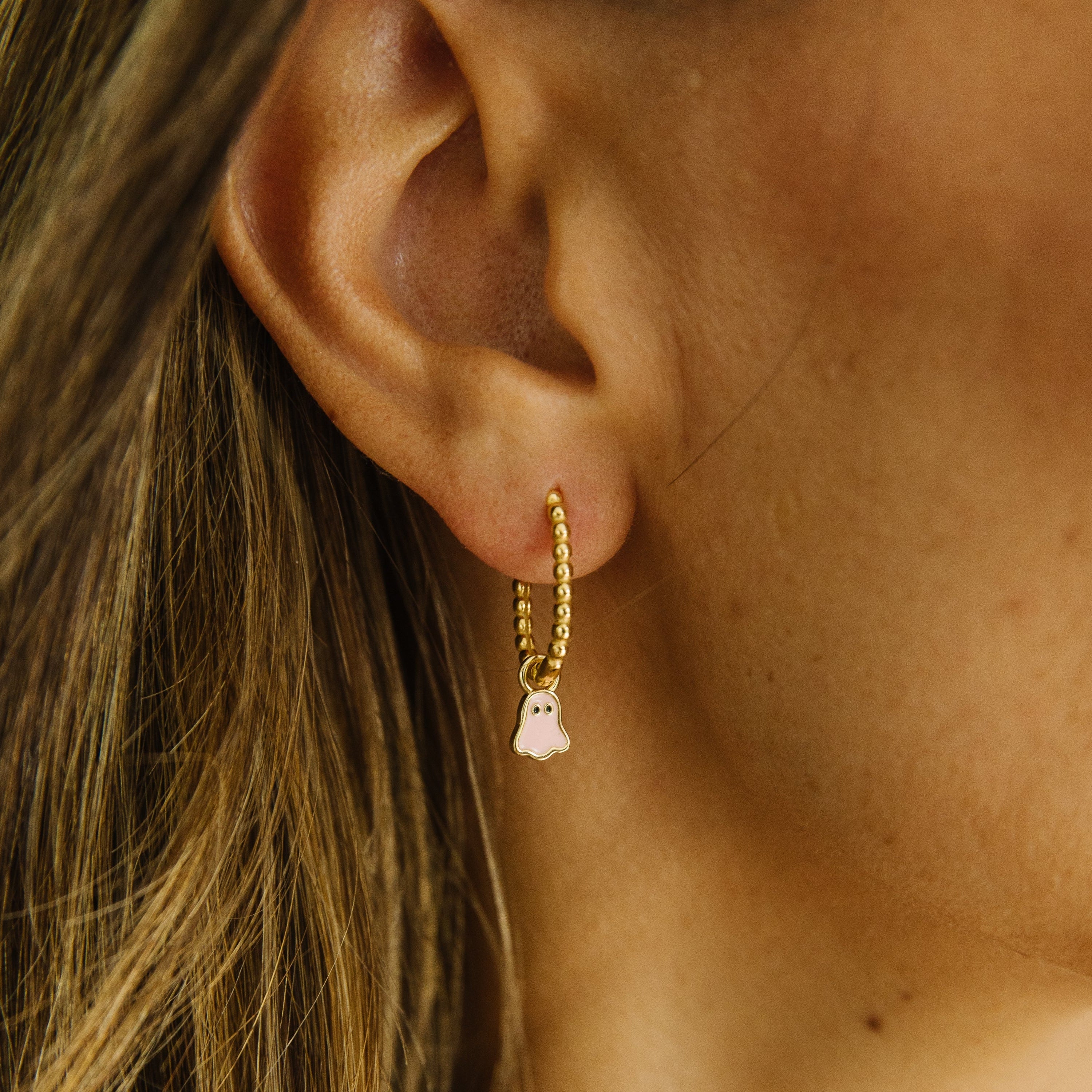 A close-up of a model's ear wearing a delicate gold huggie hoop earring with a small dangling light pink ghost charm on beaded hoop.