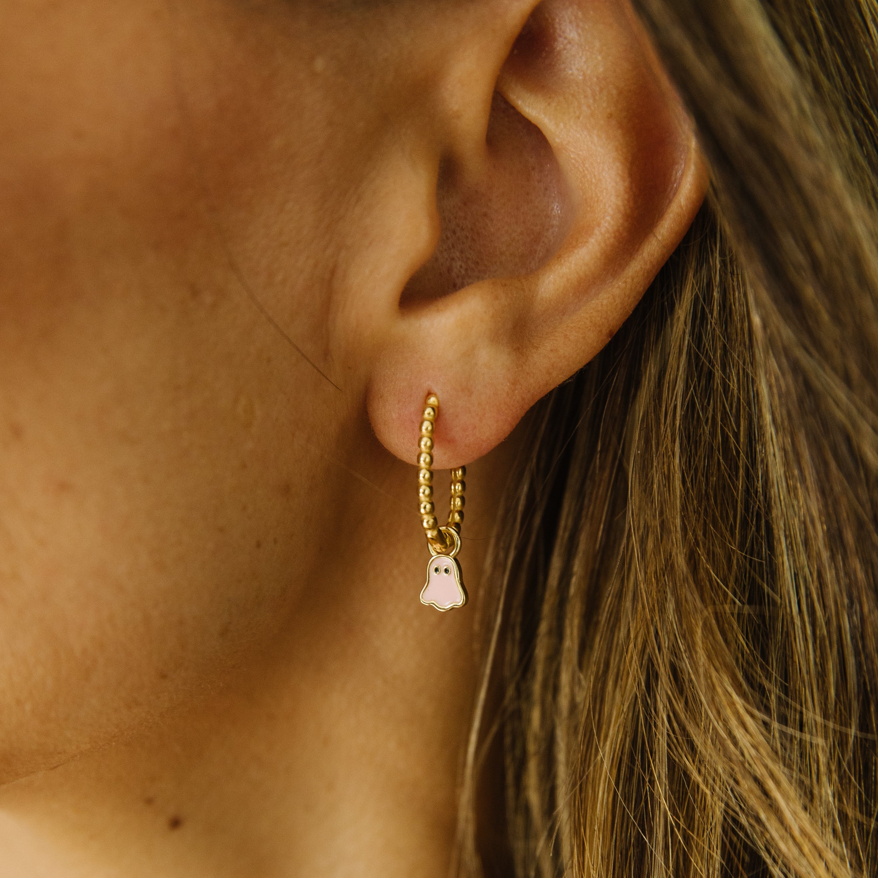 A close-up of a model's ear wearing a delicate gold huggie hoop earring with a small dangling light pink ghost charm.