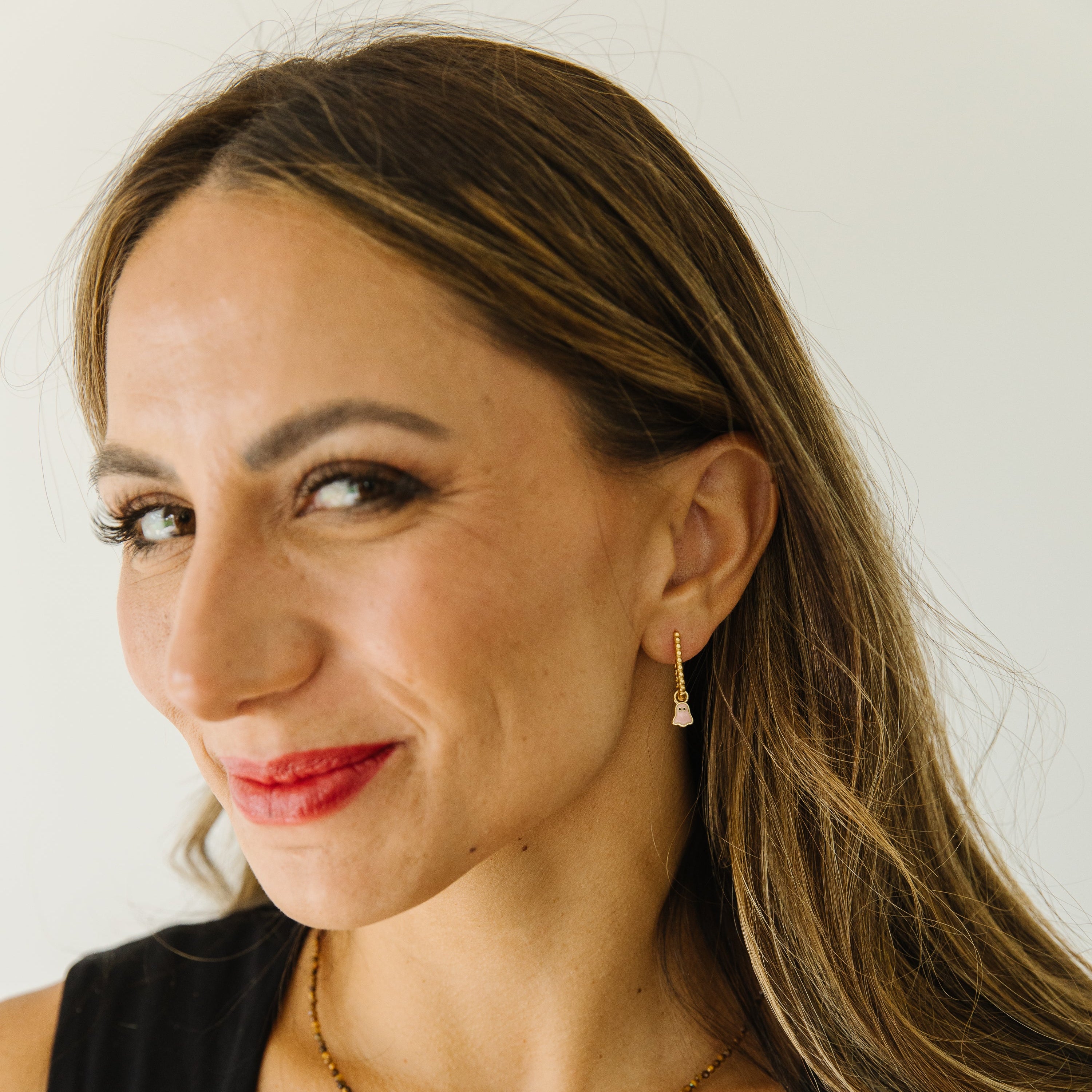 A close-up portrait of a woman with long brown hair wearing a delicate gold necklace and a small gold hoop earring with pink ghost charm, set against a plain light background.