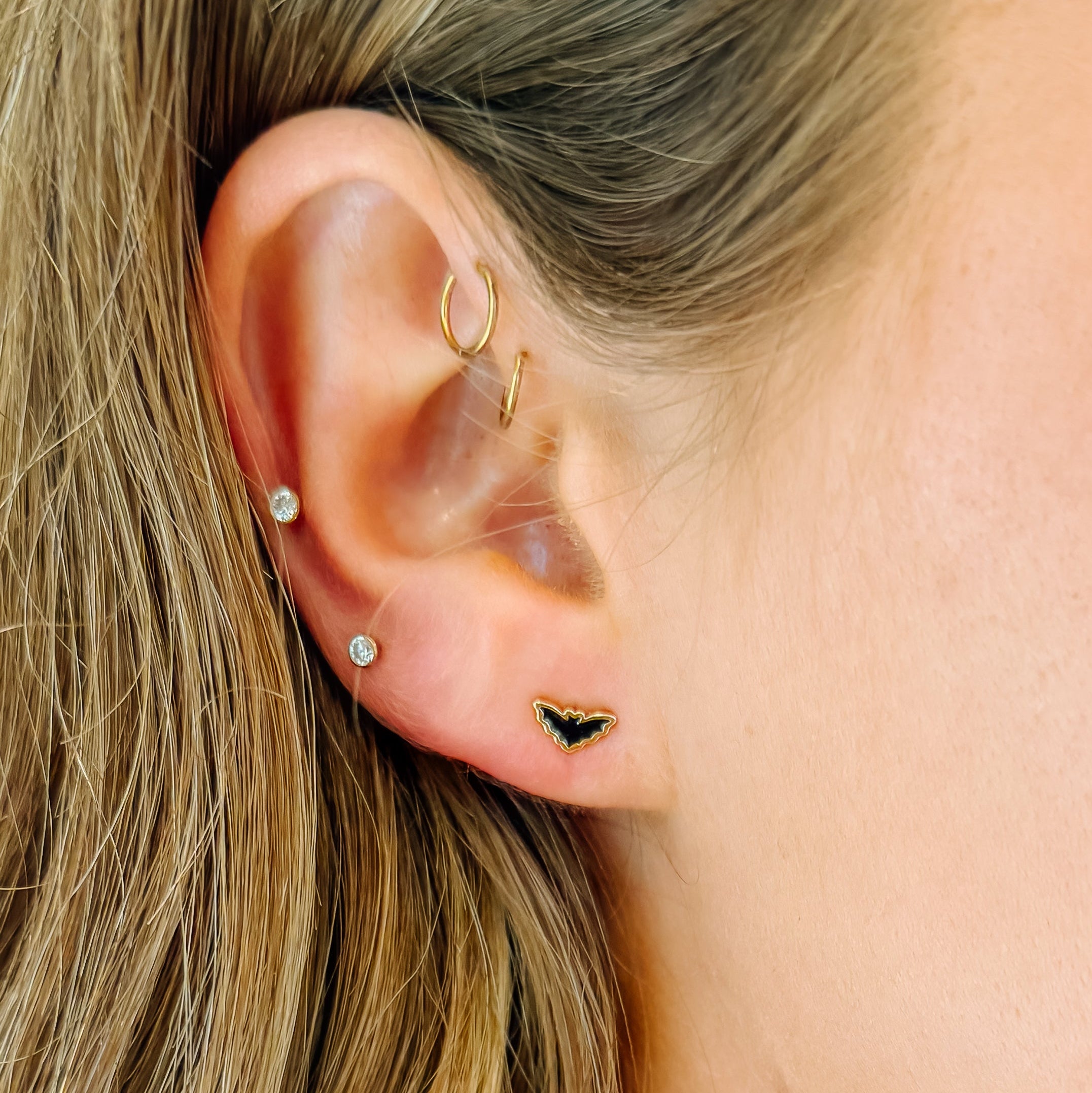Close-up of a woman's ear showcasing multiple gold piercings, including a small bat-shaped stud, a curved hoop, and two small studs with clear gemstones.
