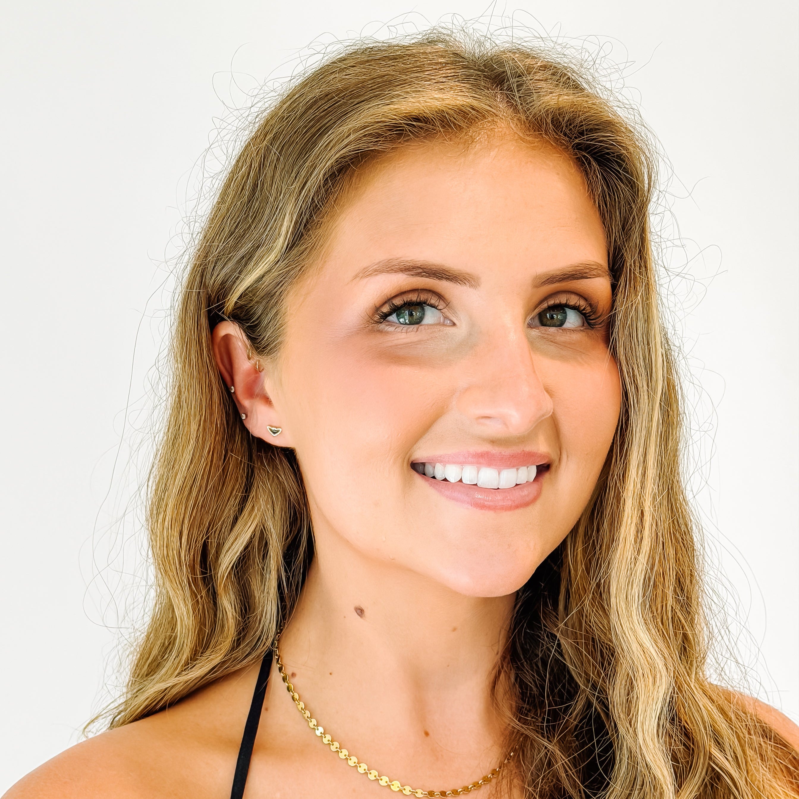 A smiling young woman with long wavy blonde hair wearing a delicate gold chain necklace, bat stud earrings and a black top, photographed against a clean white background.