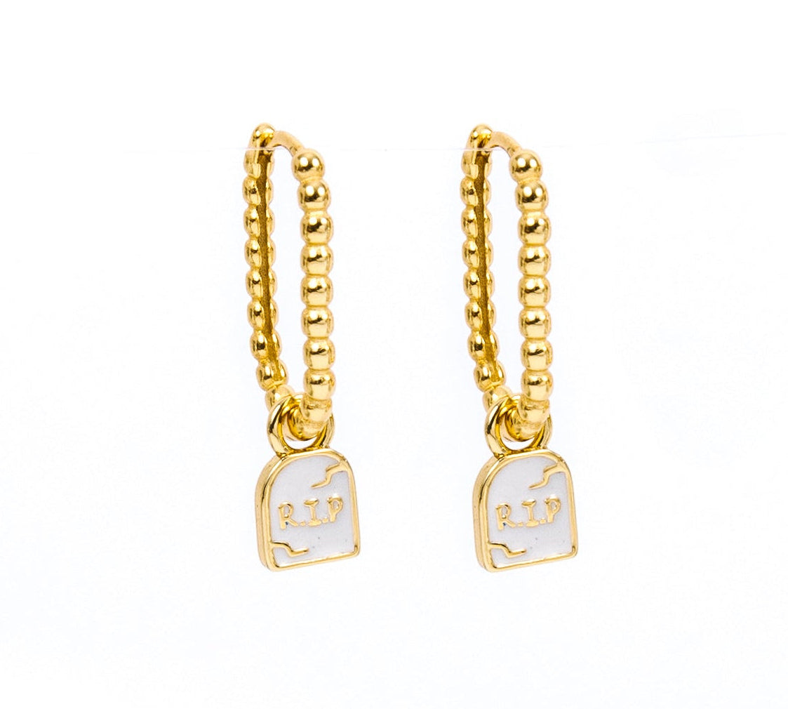 A pair of gold huggie hoop earrings with a small white enamel pendant featuring a tombstone with RIP on a plain white background.
