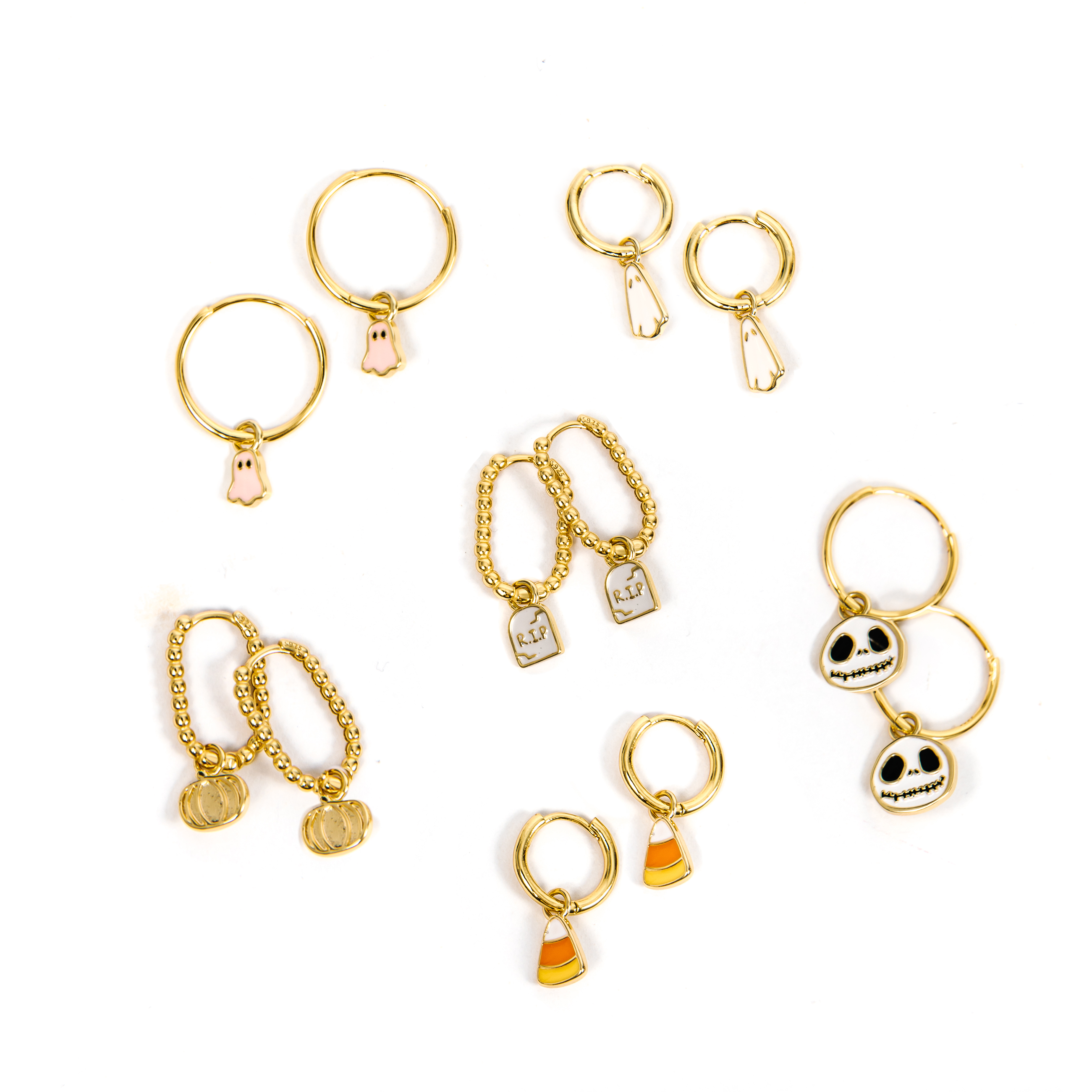 A set of gold-toned jewelry charms on hoop huggies with small decorative pendants, including a skull, a candy corn, a tombstone charm, a white ghost, a pink ghost, and a gold pumpkin, arranged on a plain white background.