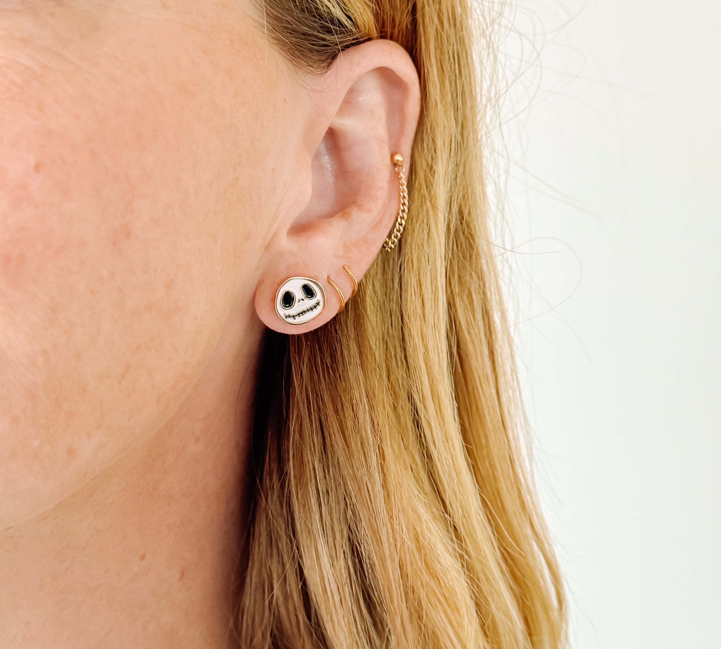 A close-up of a woman's ear wearing a small round stud earring with a black and white skull design, along with subtle gold ear cuffs, against a plain light background.
