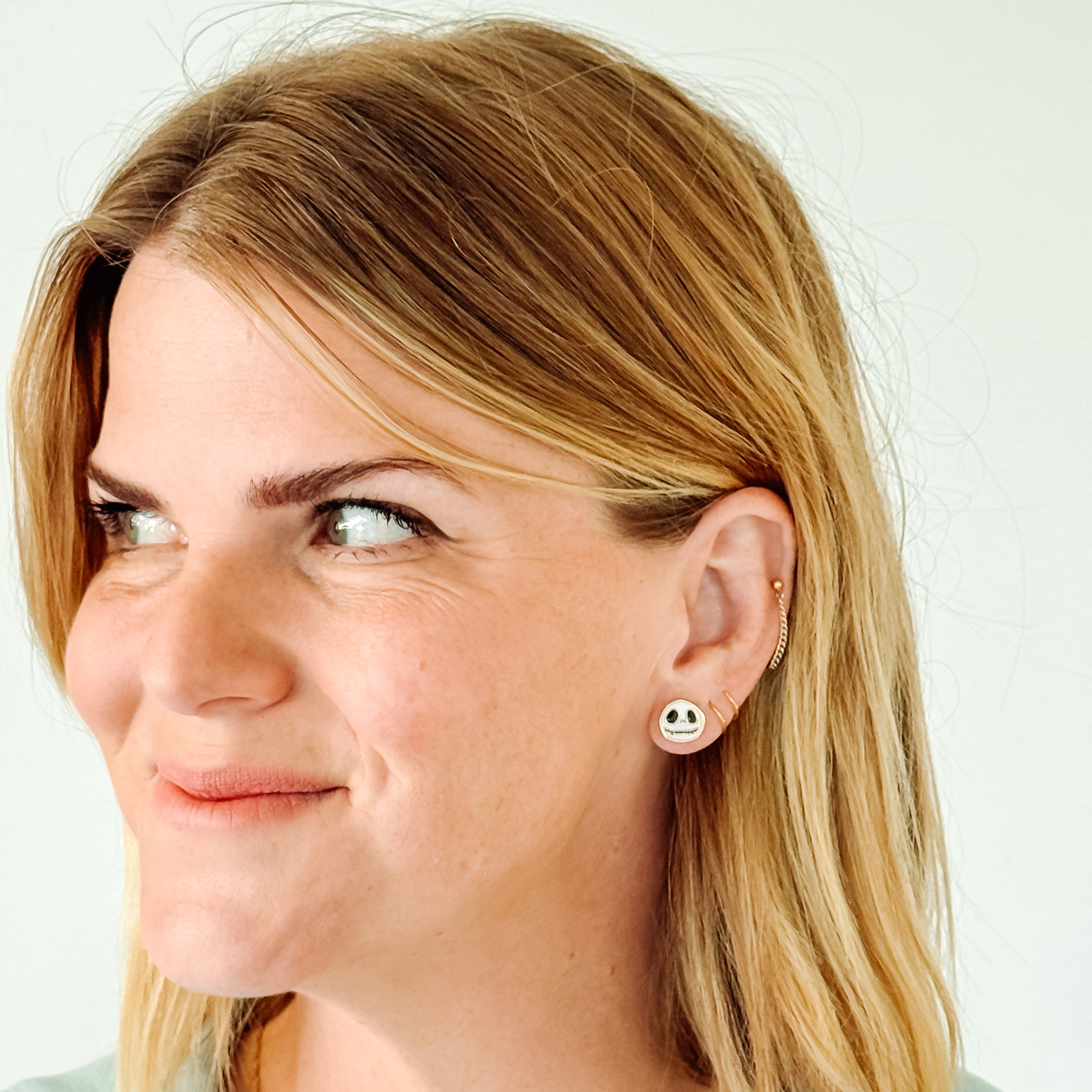 A close-up of a woman with blonde hair wearing a small white stud earring with a black smiley face, a delicate gold chain necklace, and a light blue top against a plain white background.